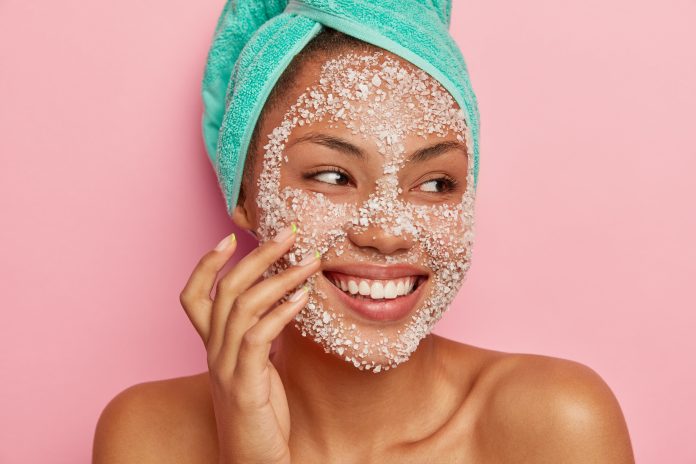 Close up portrait of glad African American woman massages cheeks, applies sea salt scrub, looks away, has gentle smile, shows white teeth, wears turquoise towel, happy to get spa treatments. peeling