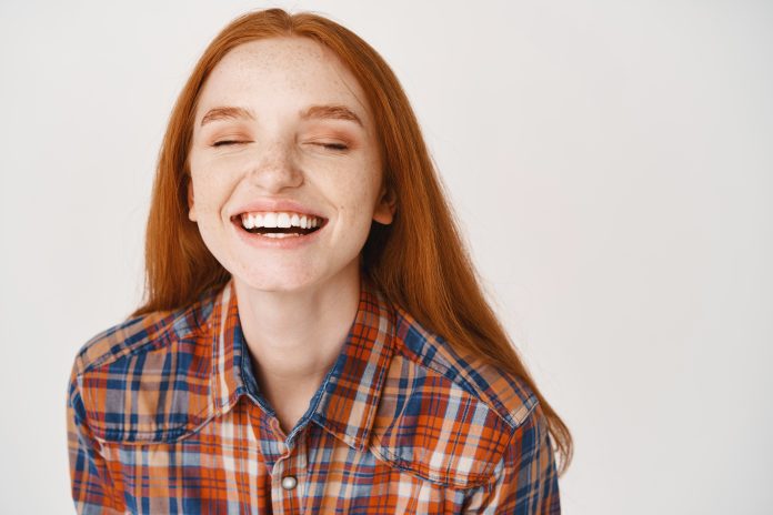 Close-up of beautiful redhead woman laughing with closed eyes, standing over white background uśmiech